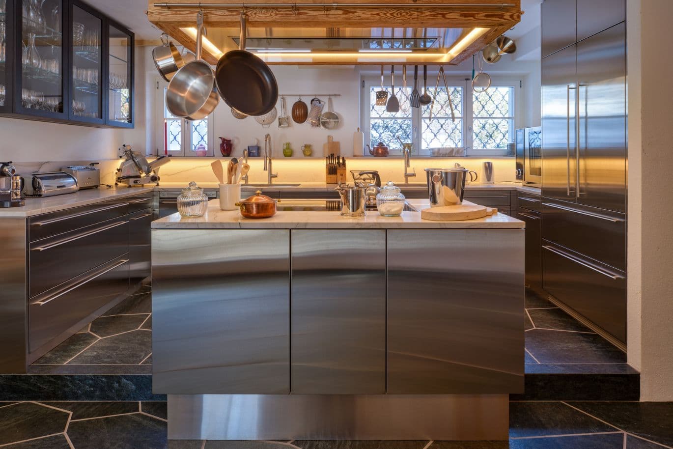Modern stainless steel kitchen with central island, marble countertop, hanging pots and pans, backlit backsplash and patterned windows.