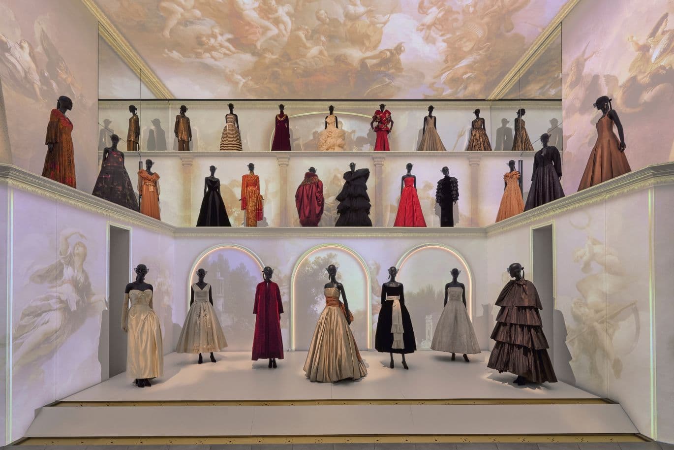 Exhibit of elegant dresses on mannequins in a three-tiered display, with ornate ceiling and archway decor.
