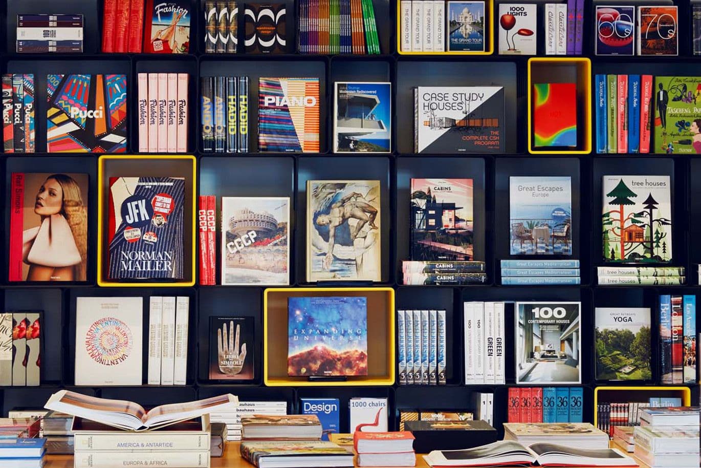 Une bibliothèque remplie de livres et de magazines colorés sur les thèmes de l'art, du design et du voyage. Au premier plan, une table avec des livres ouverts.