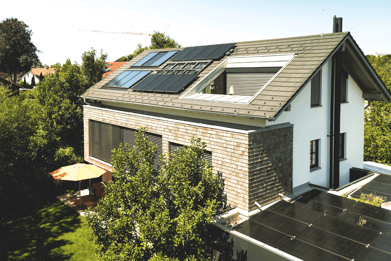 Modern house with solar panels on the roof, surrounded by lush greenery and a patio with an orange umbrella in the backyard.