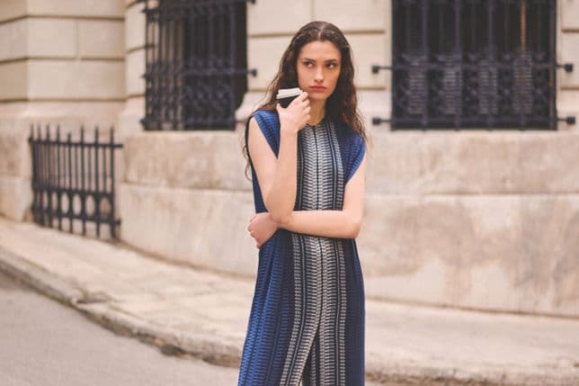 Eine Frau in einem blau gestreiften Kleid steht mit einer Kaffeetasse in der Hand vor einem Steingebäude mit schwarzen Eisenfenstern.