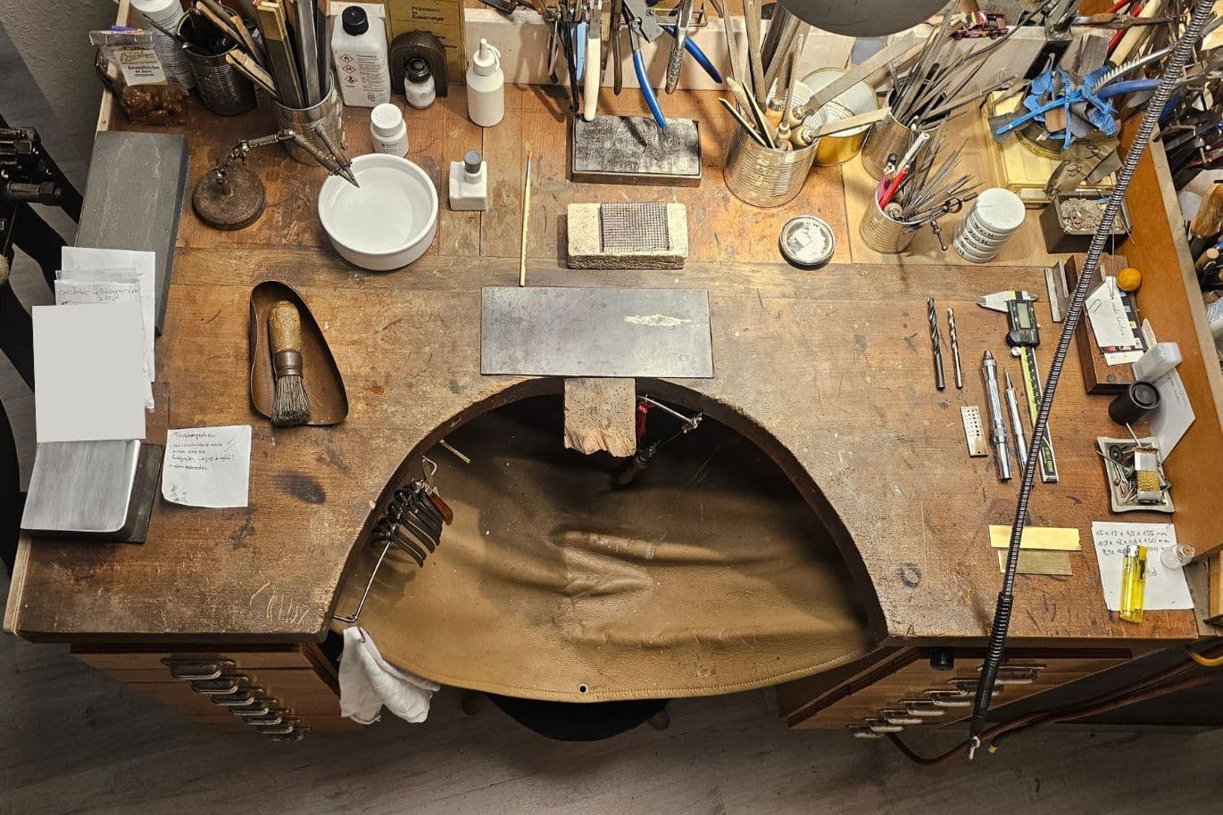 Überfüllte Werkbank mit Werkzeugen, Notizblock, Messschiebern, Behältern und einer Lederschürze auf einer Holzoberfläche.