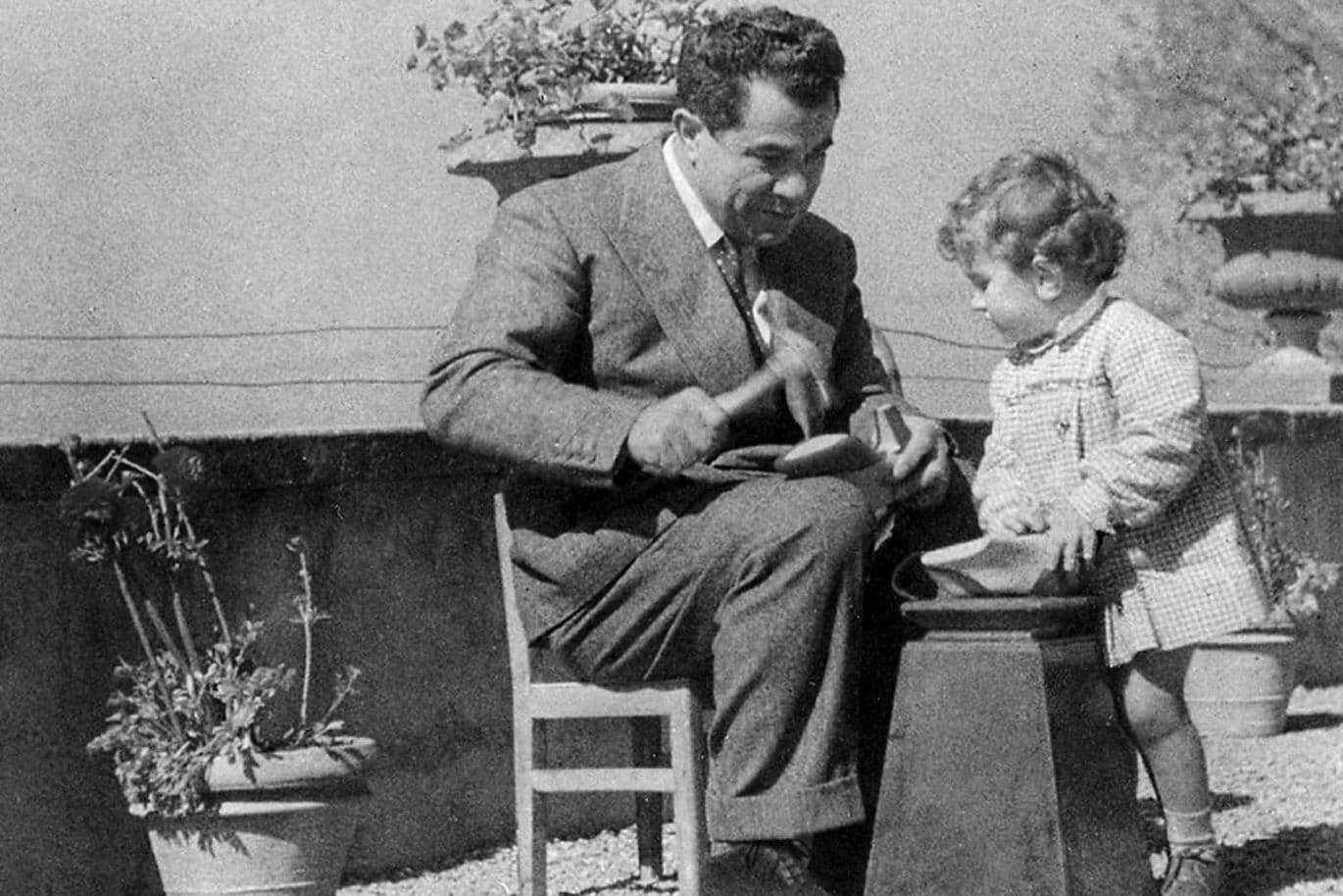A man in a suit sits outdoors on a chair playing with a small child wearing a coat. They are surrounded by potted plants on a gravel floor.