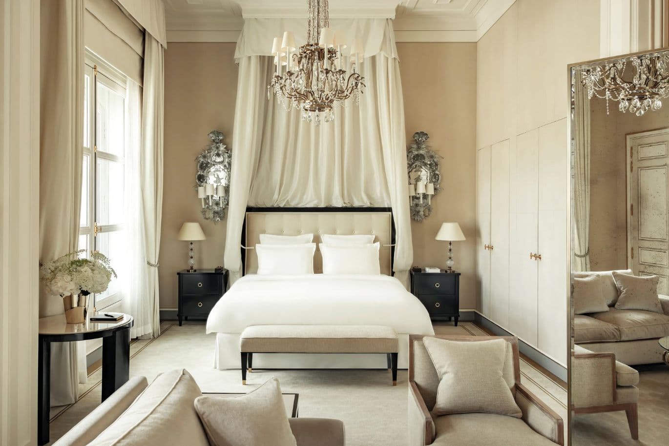 Elegant bedroom with a luxurious canopy bed, chandeliers, and beige tones. Features plush seating, mirrored wall, and soft lighting.