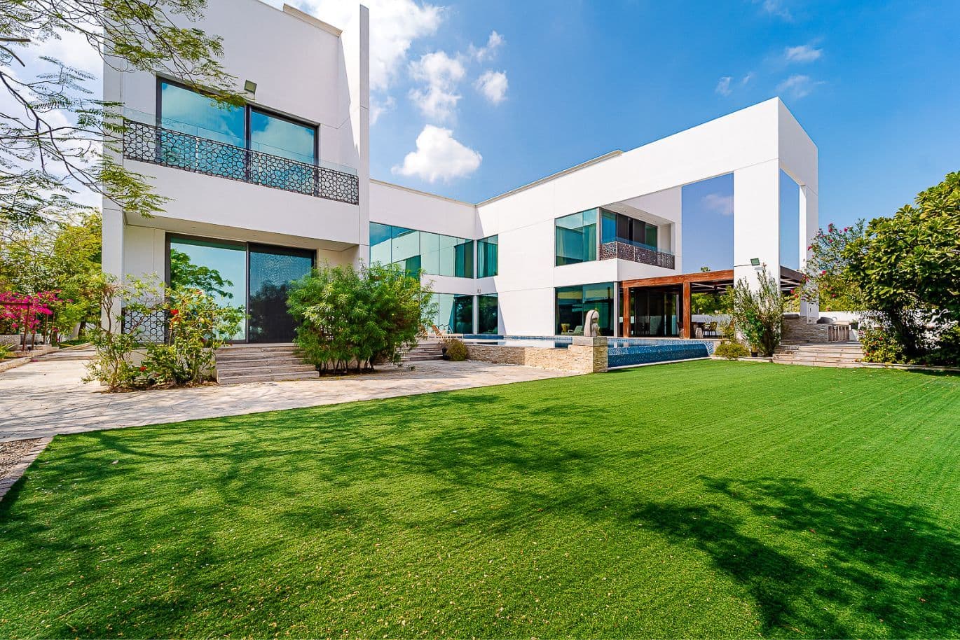 Modern white house with large windows, a spacious green lawn, and a pool, surrounded by trees under a clear blue sky.