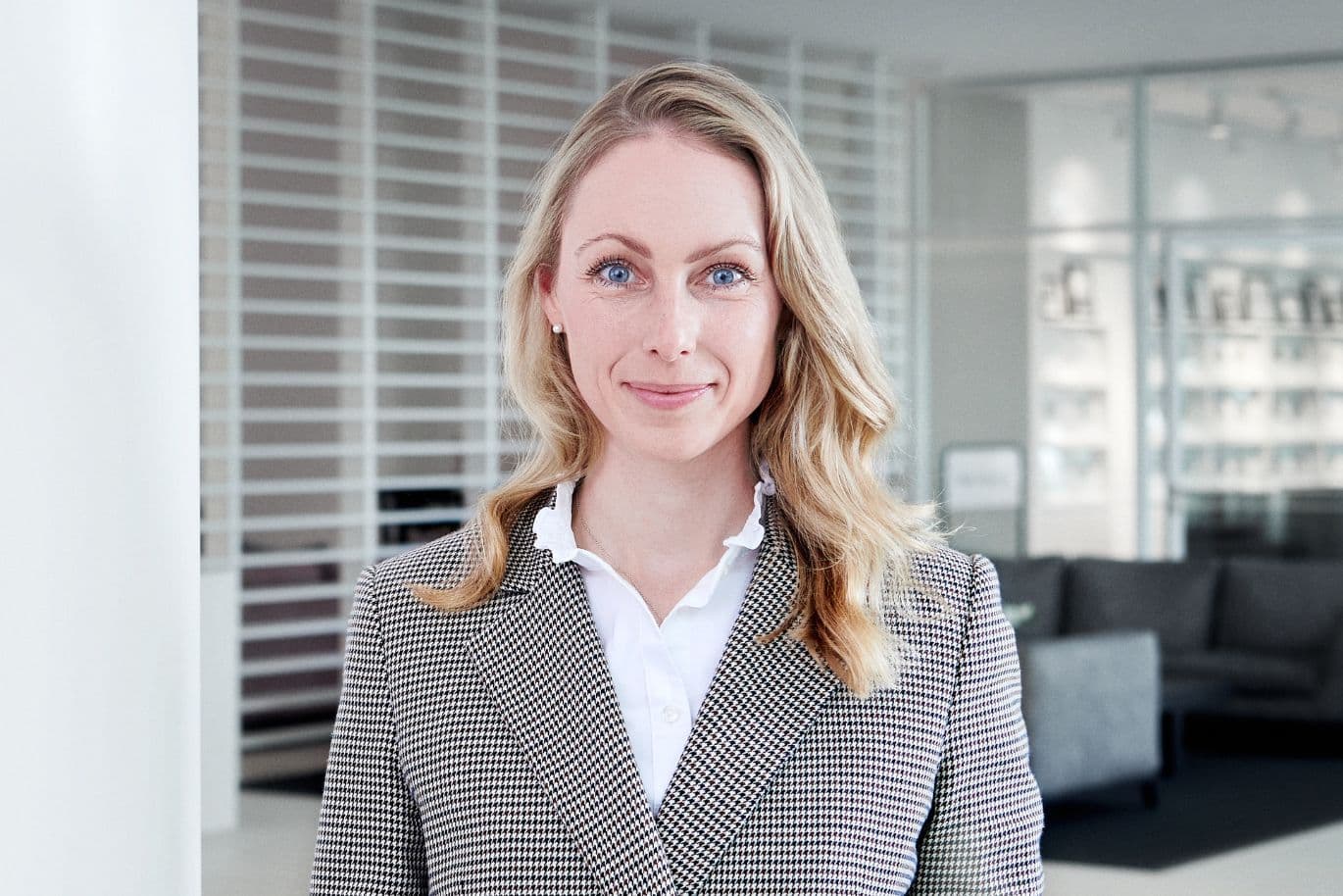 Eine Frau mit langen blonden Haaren, die einen karierten Blazer und eine weiße Bluse trägt, steht mit selbstbewusstem Gesichtsausdruck in einer modernen Büroumgebung.