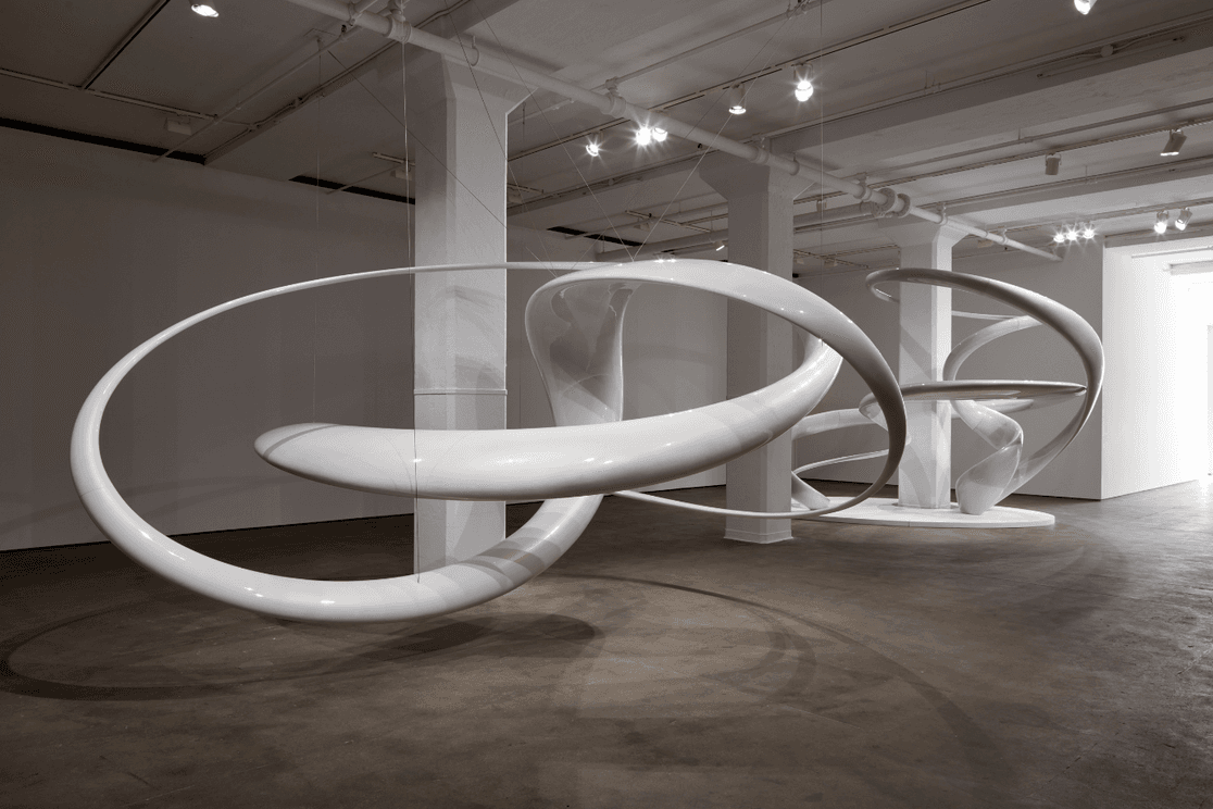 Eine moderne Kunstinstallation aus großen, ineinander verschlungenen weißen Schleifen, die in einem Galerieraum aufgehängt sind, wobei die Decke mit Strahlern beleuchtet wird.