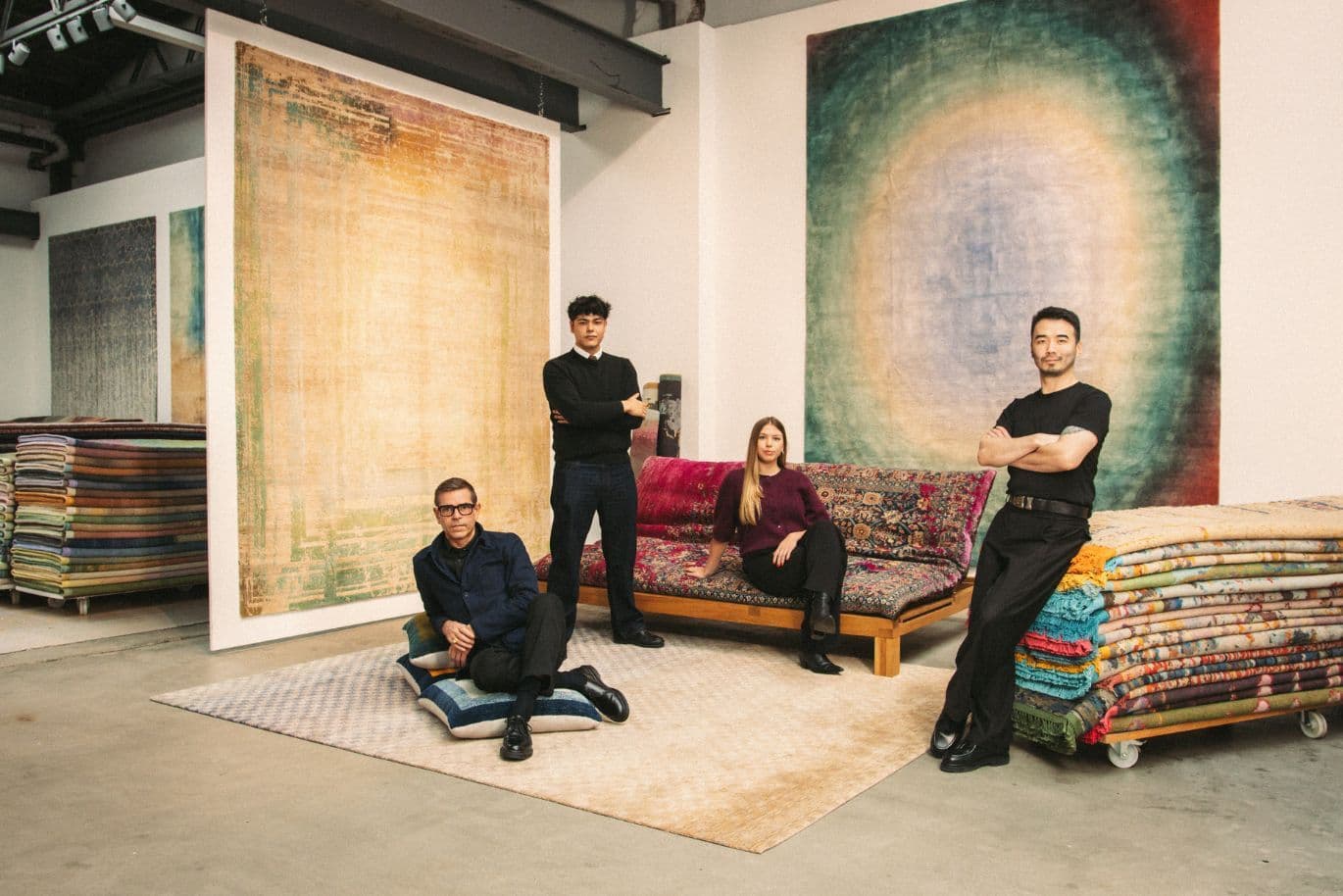 Vier Personen posieren selbstbewusst in einem Raum, in dem bunte Teppiche hängen und um sie herum gestapelt sind. Zwei stehen, während zwei auf einem gemusterten Sofa sitzen.