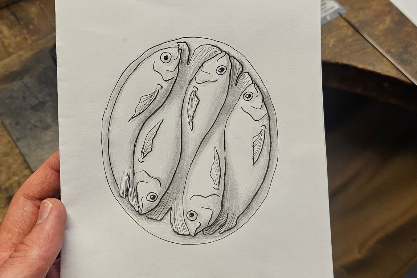 Hand, die eine Skizze von vier symmetrisch angeordneten Fischen hält, mit Bleistift auf weißem Papier gezeichnet.