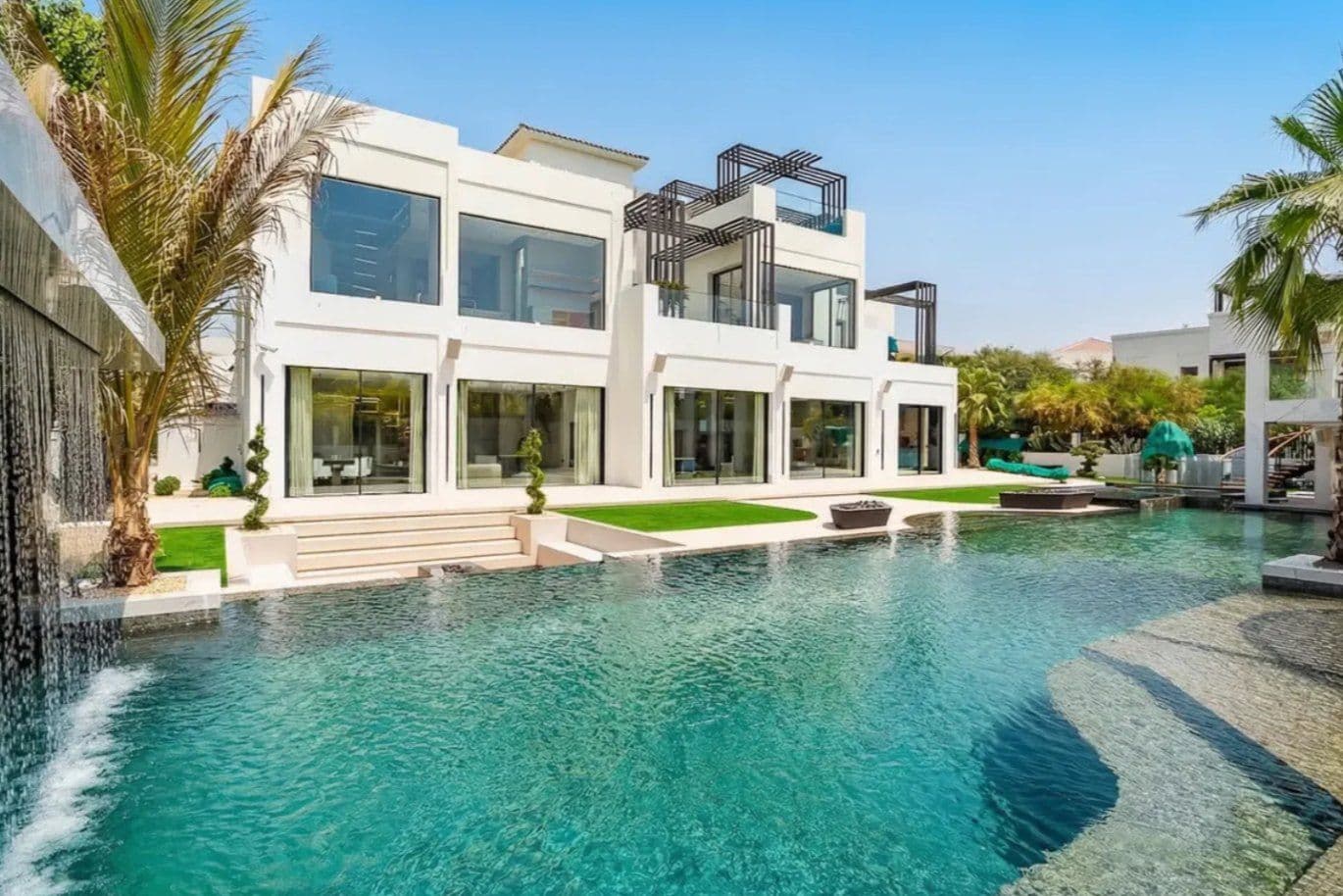 Two-story white villa with floor-to-ceiling windows, a turquoise pool, green lawn, and palm trees under a clear blue sky.