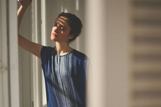 Eine Person in einem blau gemusterten Kleid lehnt an einer Wand, die Augen geschlossen, während das Sonnenlicht Schatten auf ihr Gesicht wirft.