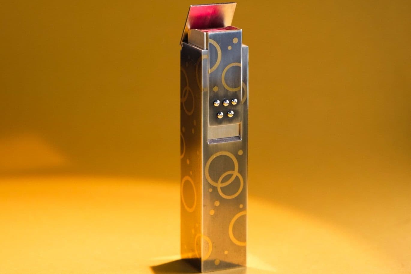 Ein metallischer, rechteckiger Lippenstift mit kreisförmigen Mustern und einem teilweise geöffneten Deckel steht vor einem warmen, gelb-orangefarbenen Hintergrund.