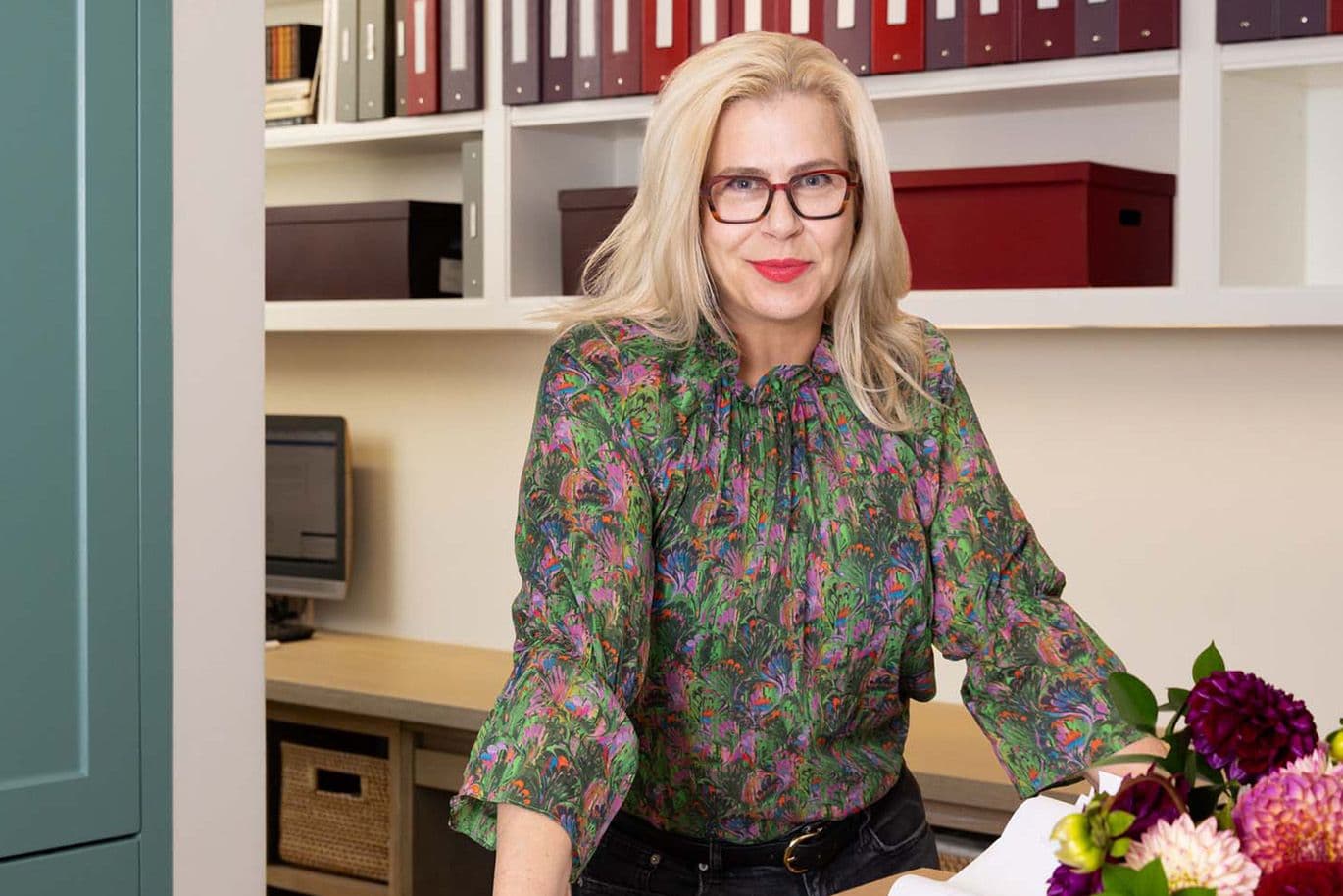 Woman wearing glasses and a floral blouse smiles at a desk surrounded by colourful files and flowers.