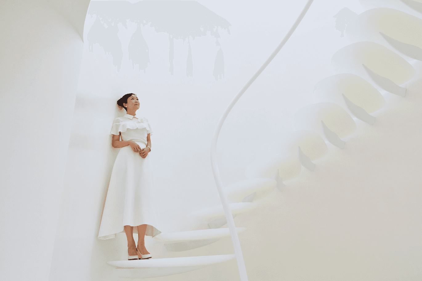 Woman in a white dress and heels stands on a white spiral staircase, inside a bright, minimalist curved-wall interior.