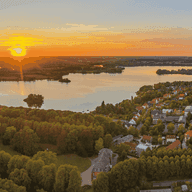 Luftaufnahme eines Sonnenuntergangs über einem ruhigen See, umgeben von üppigem Grün und einem kleinen Dorf, mit leuchtenden Orange- und Blautönen am Himmel