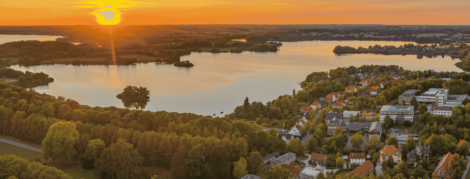 Luftaufnahme eines Sonnenuntergangs über einem ruhigen See, umgeben von üppigem Grün und einem kleinen Dorf, mit leuchtenden Orange- und Blautönen am Himmel