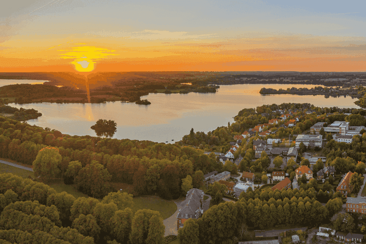 Luftaufnahme eines Sonnenuntergangs über einem ruhigen See, umgeben von üppigem Grün und einem kleinen Dorf, mit leuchtenden Orange- und Blautönen am Himmel