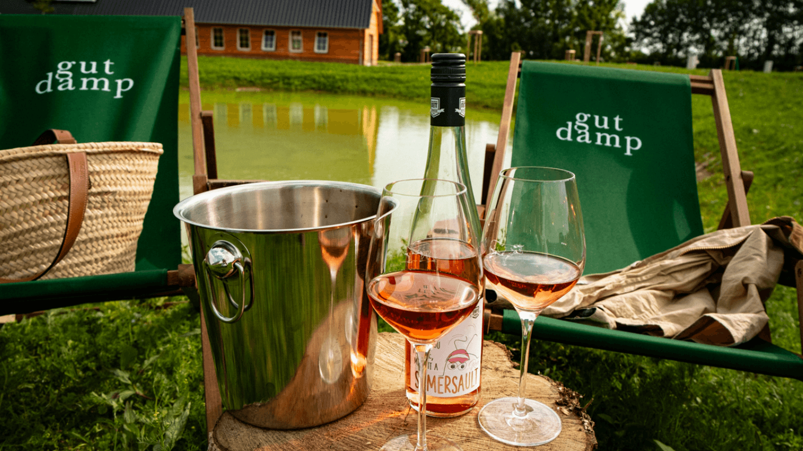 Two glasses of rosé wine and a bottle on a tree stump, with a metal ice bucket nearby. Green chairs and a pond are in the background.