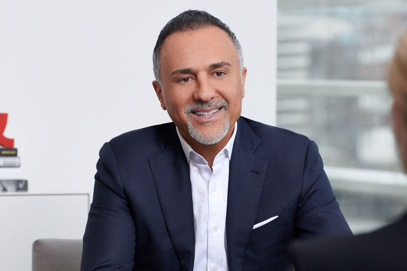 Smiling man in a navy suit sits at a table, with blurred background and books visible on the left.
