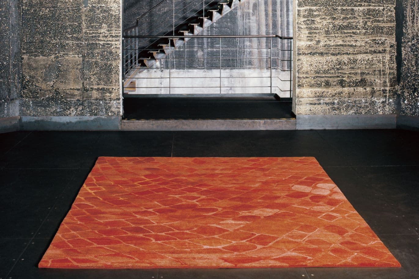Roter gemusterter Teppich auf einem dunklen Boden, vor einer Betonwand mit einer Metalltreppe und einem Geländer im Hintergrund.