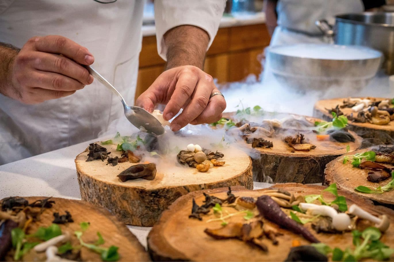 Chef plating gourmet dishes with mushrooms and greens on wooden slabs, surrounded by mist from dry ice.