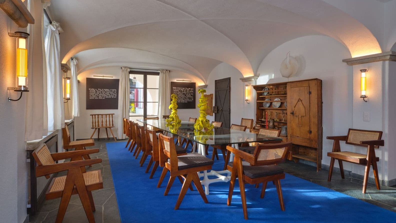Sonnenbeschienener Speisesaal mit Gewölbedecke, blauem Teppich, langem Glastisch, Holzstühlen und zwei gelben skulpturalen Tischdekorationen.