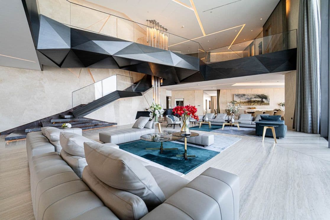 Spacious modern living room with a grand staircase, beige sofas, red and white floral arrangements, and contemporary decor.