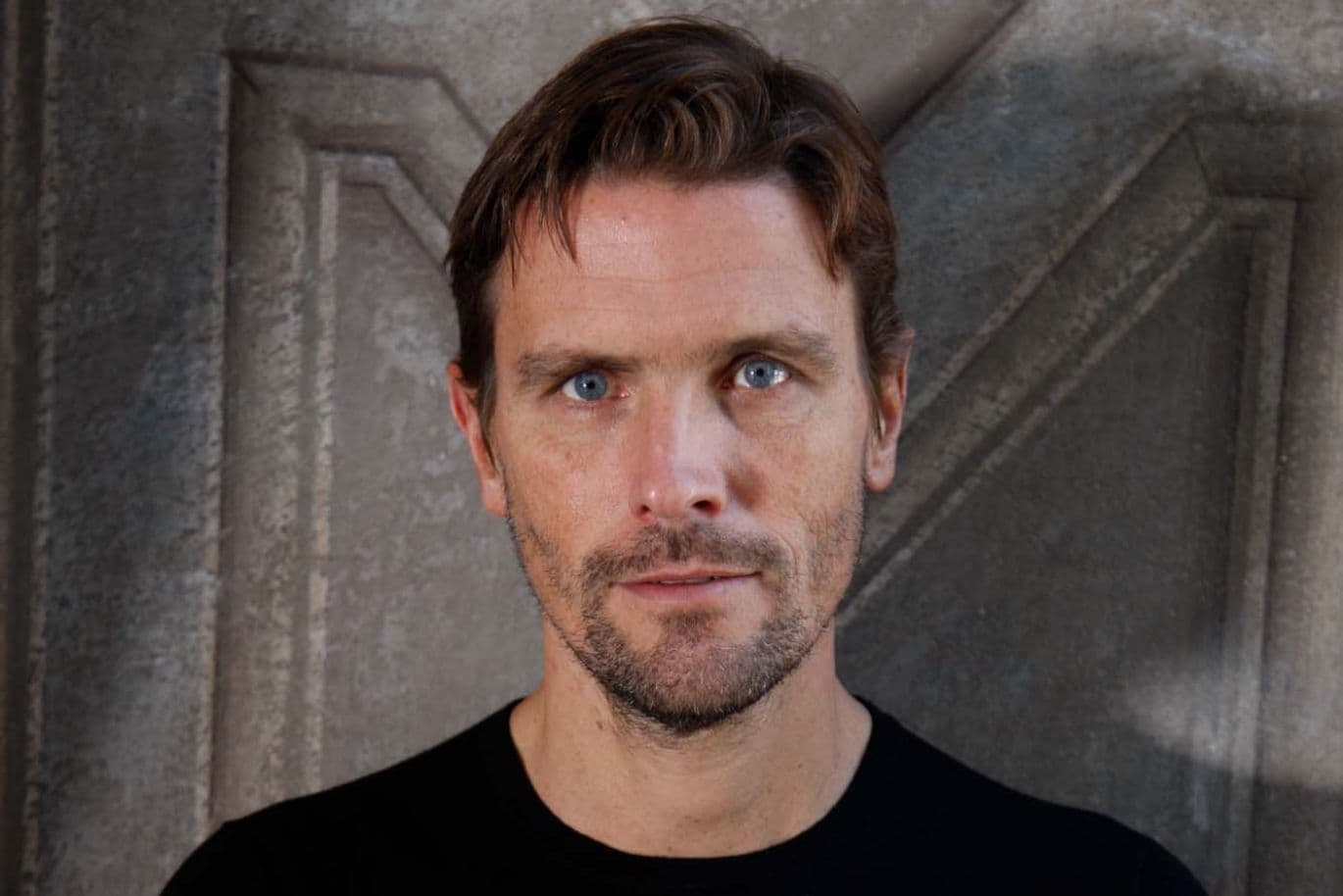 A man with short hair and a beard stands with his arms crossed in front of a textured grey background, wearing a black jumper.