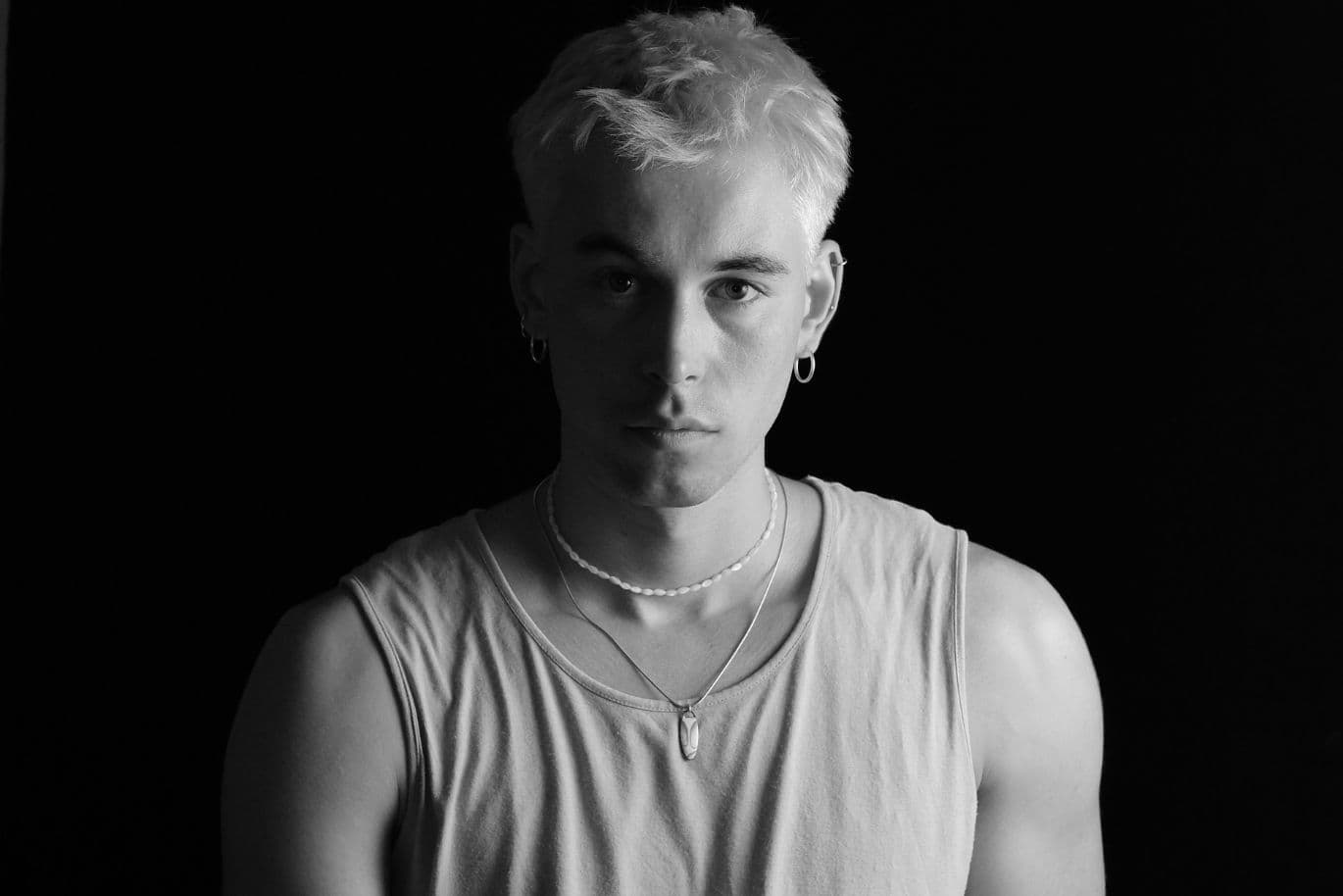 Schwarz-Weiß-Porträt einer Person mit kurzen, hellen Haaren, die ein ärmelloses Oberteil, Halsketten und Ohrringe trägt, vor einem dunklen Hintergrund.