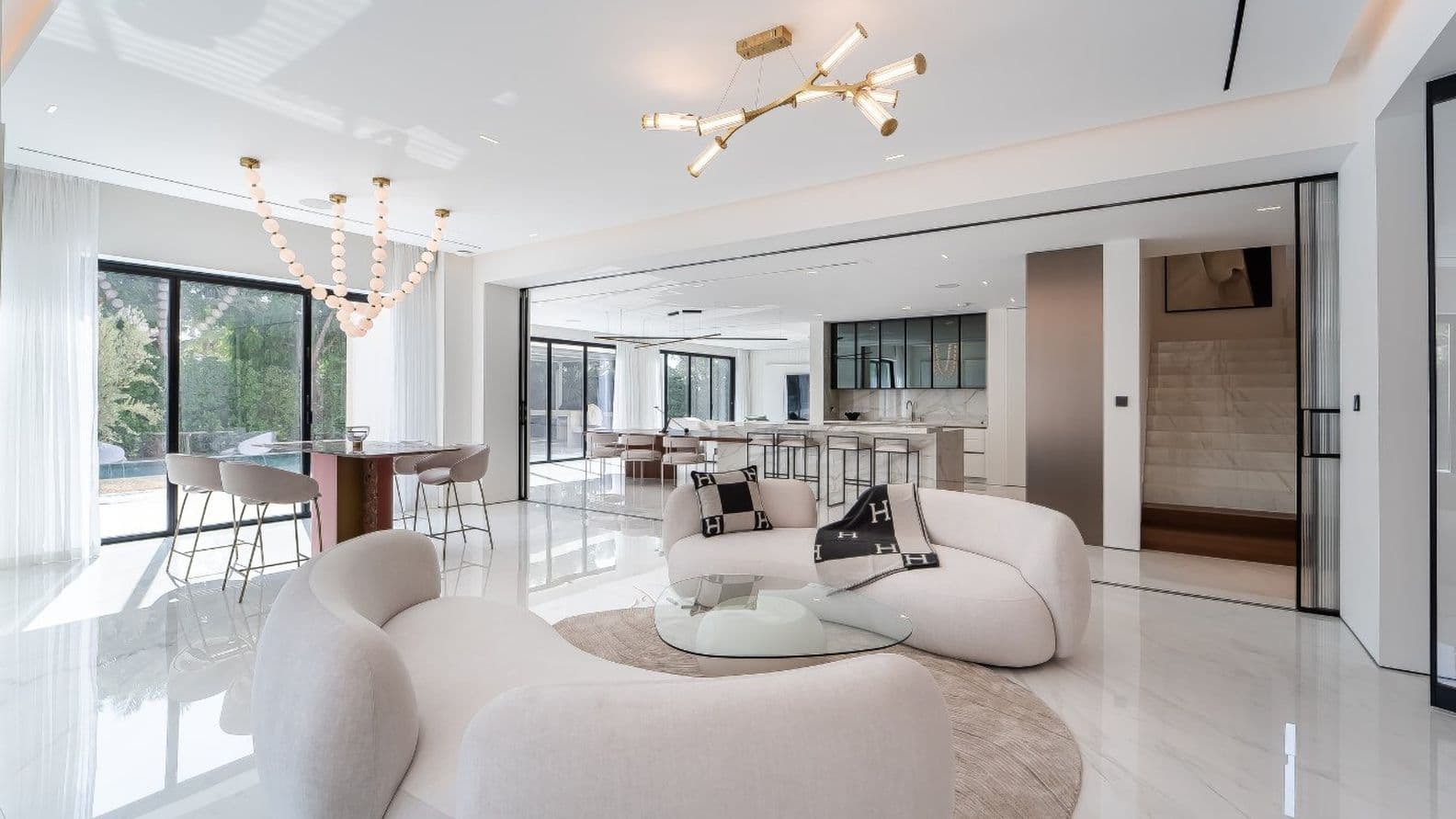 Modern open-plan living area with curved white sofas, glass coffee table, two chandeliers, and bar stools at a kitchen island. Large windows provide light.