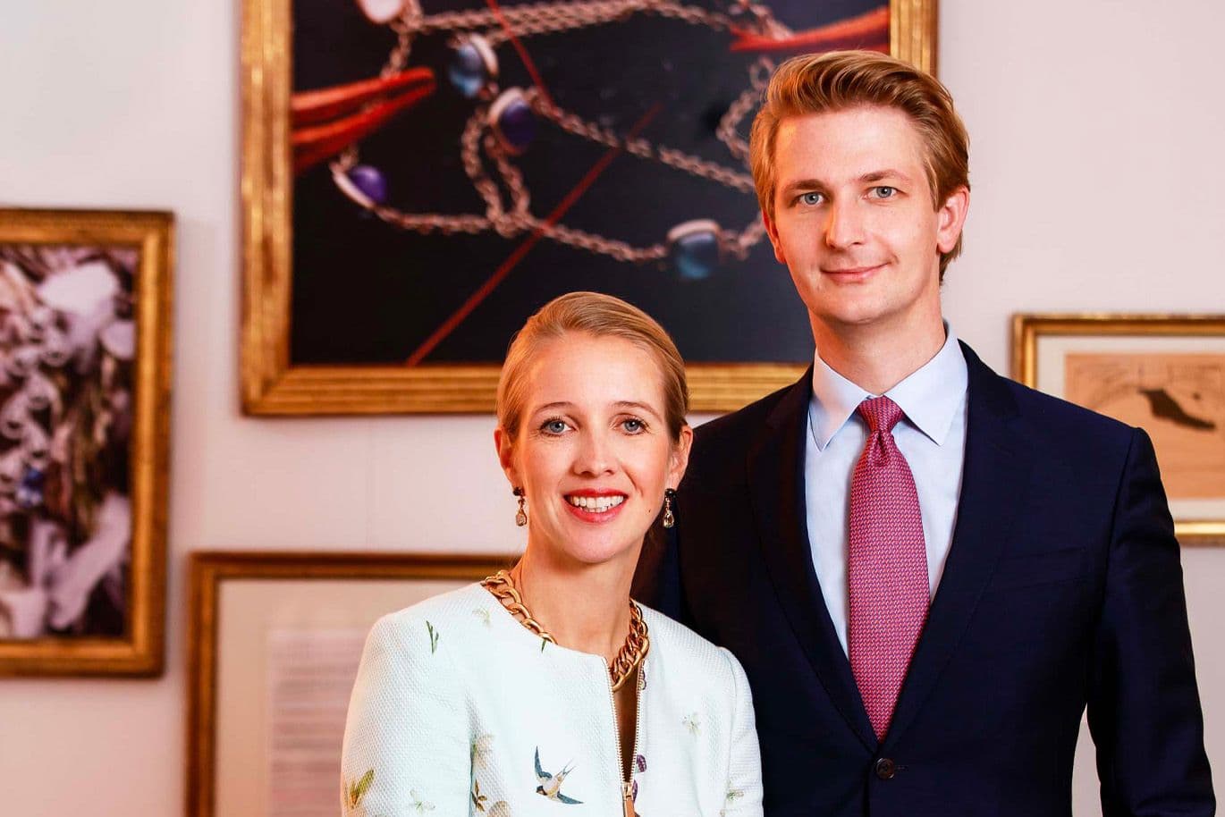 A smiling man in a suit and a woman in a floral jacket stand in front of framed artwork.