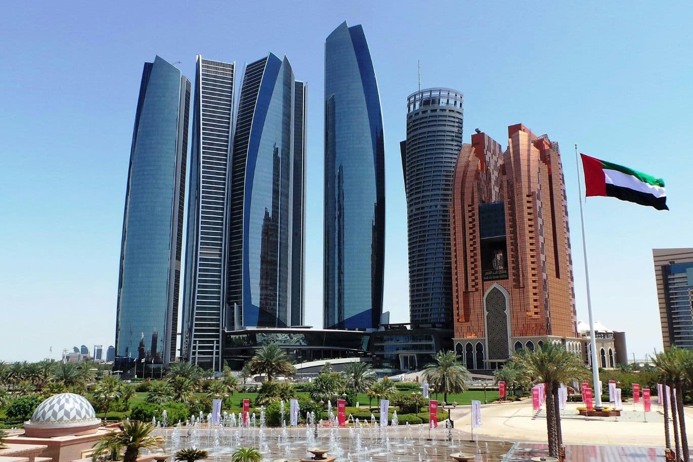 Skyline of Abu Dhabi featuring modern skyscrapers, including Etihad Towers, with a UAE flag in the foreground and a clear blue sky.