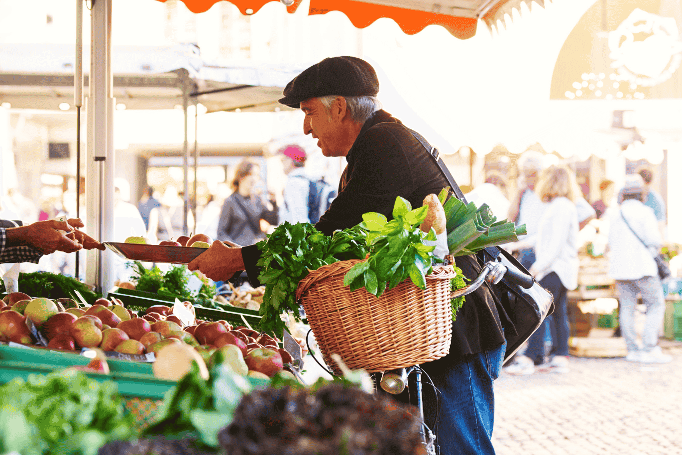 Elderly man with a basket of greens shopping at an outdoor market stall filled with fresh produce.