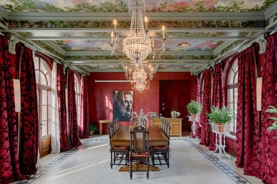 Opulent dining room with ornate chandeliers, red floral drapes, art on walls, and a long wooden table with decorative chairs.