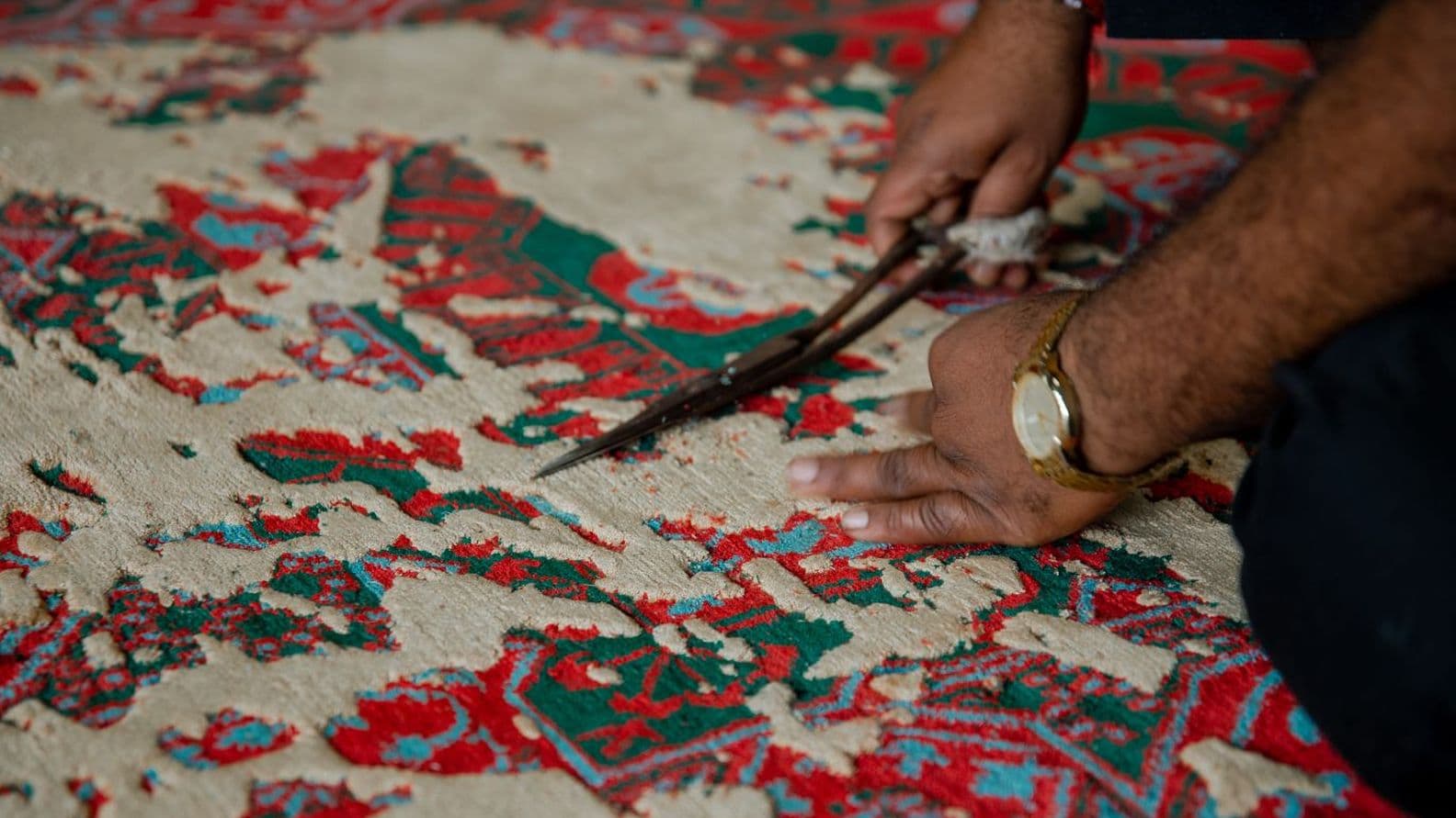 Hände, die mit einer Schere einen bunten, aufwendig gemusterten Teppich zuschneiden. Der Teppich hat rote und grüne Muster auf beigem Hintergrund.