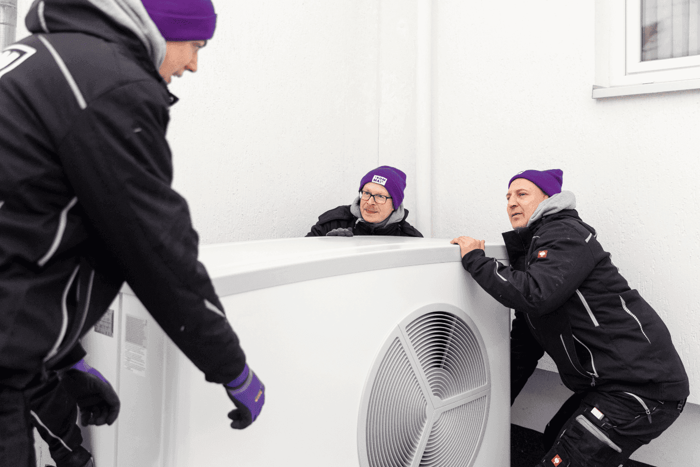 Three people in winter clothing and purple hats are installing a large white HVAC unit outside a building.