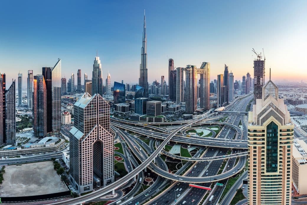 Aerial view of a modern cityscape with skyscrapers, including a tall iconic tower, and intricate highway interchanges at sunset.