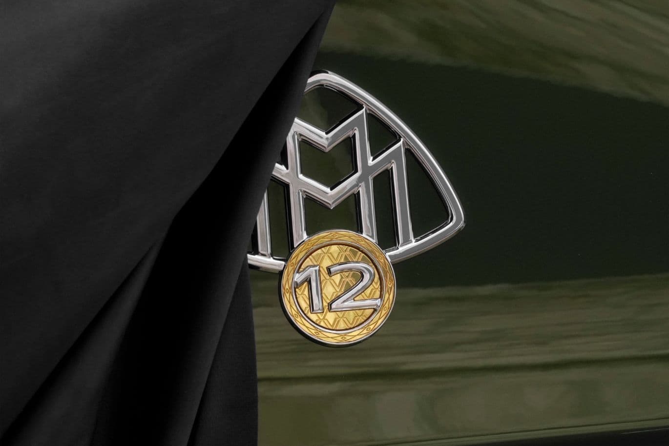 Nahaufnahme eines Luxusauto-Emblems, das teilweise von einem schwarzen Tuch verdeckt ist, mit einem metallischen Logo mit einer „12” auf einem goldfarbenen Kreis.