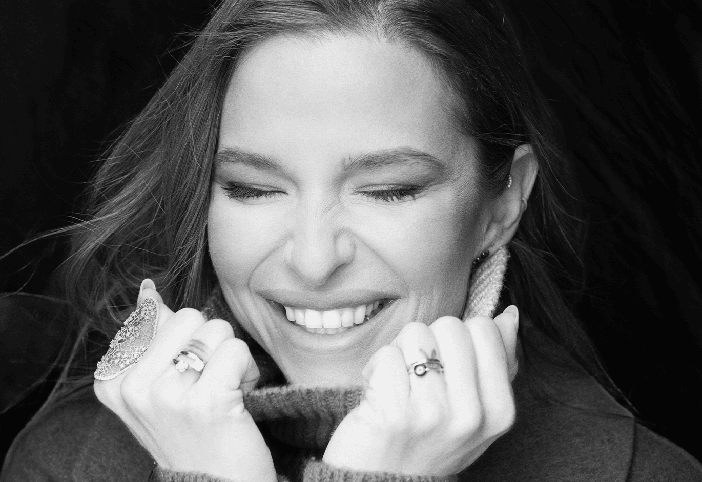 Ασπρόμαυρο πορτρέτο γυναίκας που χαμογελά κρατώντας το ζιβάγκο της, με εμφανή κοσμήματα.