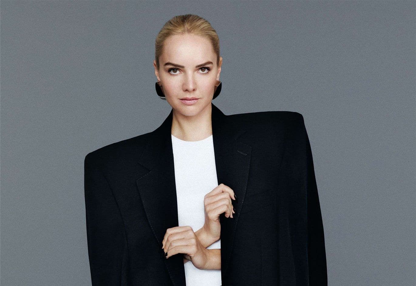 Γυναίκα με πίσω δεμένα ξανθά μαλλιά, φοράει υπερβολικά φαρδύ μαύρο σακάκι με έντονους ώμους πάνω από λευκή μπλούζα, μπροστά σε γκρι φόντο.