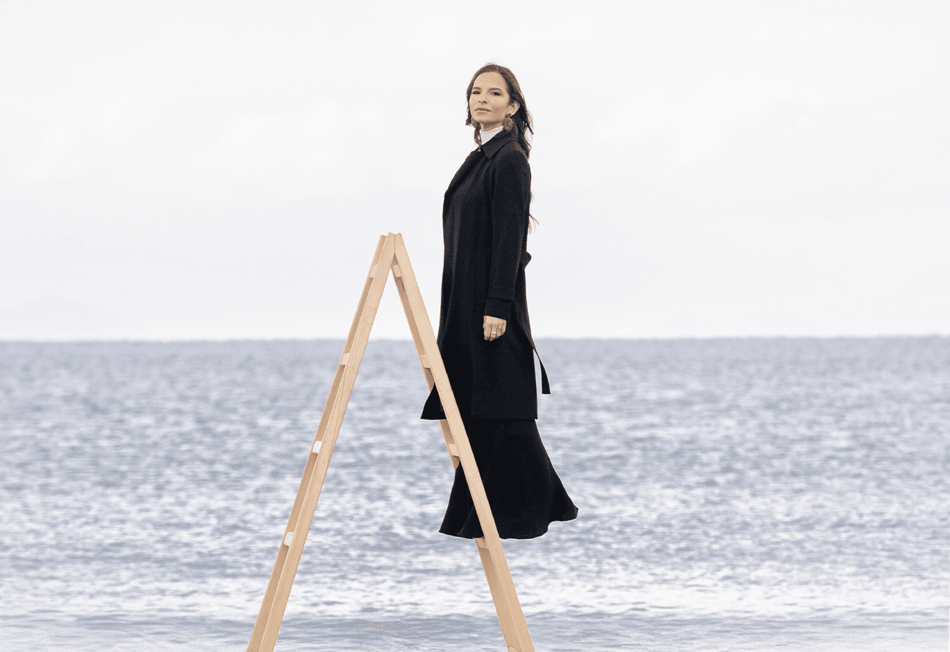 Γυναίκα με κομψό μαύρο παλτό περπατά πάνω σε ξύλινη σκάλα μέσα στη θάλασσα με φόντο τον ορίζοντα.