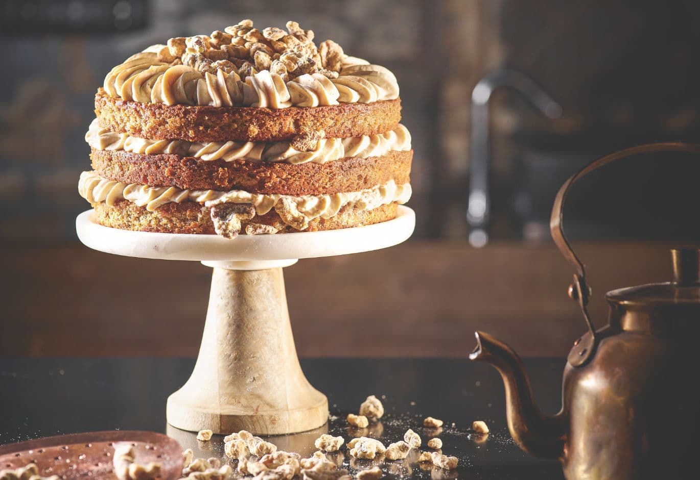 Artisanal layered cake with cream frosting and caramelized nuts on a pedestal stand, styled in a rustic kitchen setting.