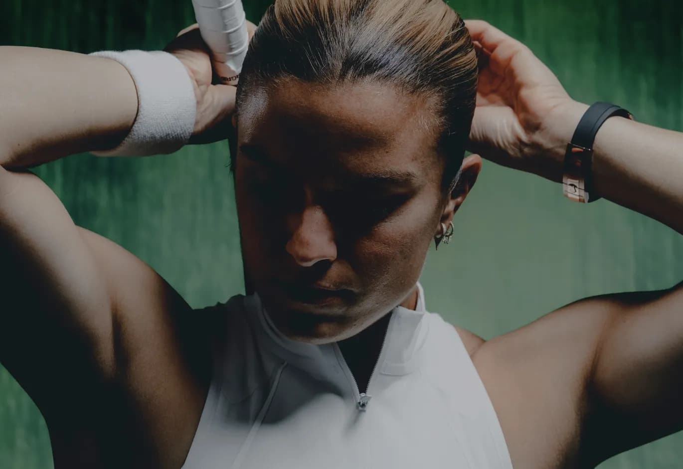 Close-up dramatic portrait of female tennis player Maria Sakkari adjusting her hair and grip before a match, set against a dark green backdrop.