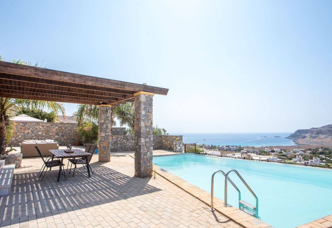 Private infinity pool with shaded pergola seating area, overlooking the sea and coastal landscape under a clear sky.