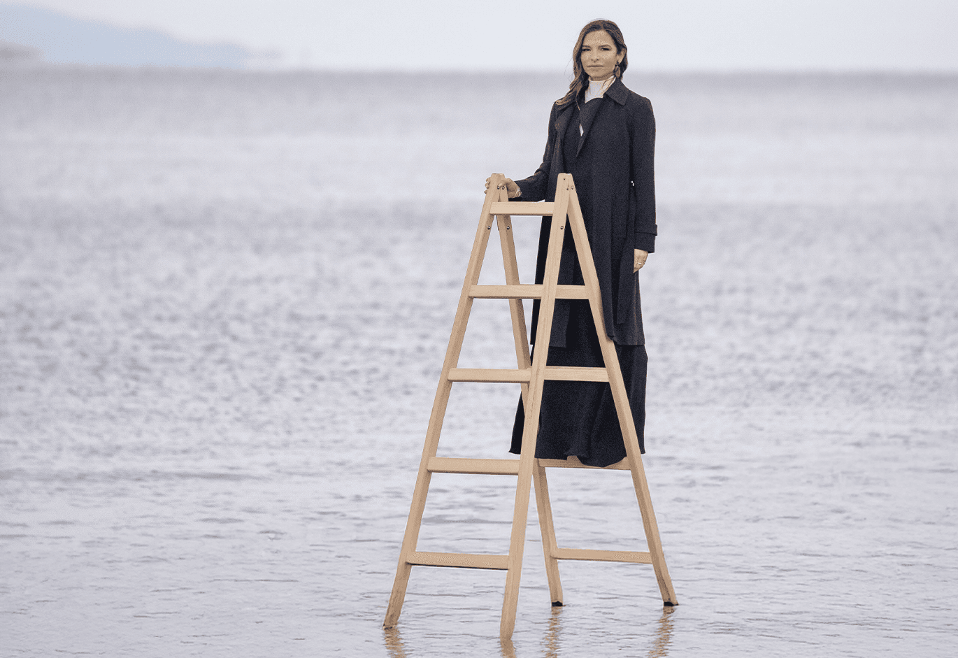 Γυναίκα με μαύρο παλτό στέκεται σε ξύλινη σκάλα μέσα στη θάλασσα, σε μινιμαλιστικό θαλάσσιο τοπίο.