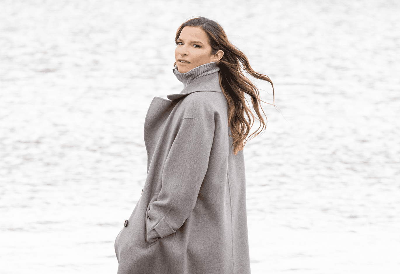 Woman in a gray coat stands by a body of water, looking over her shoulder with flowing hair, against a serene, blurred background.