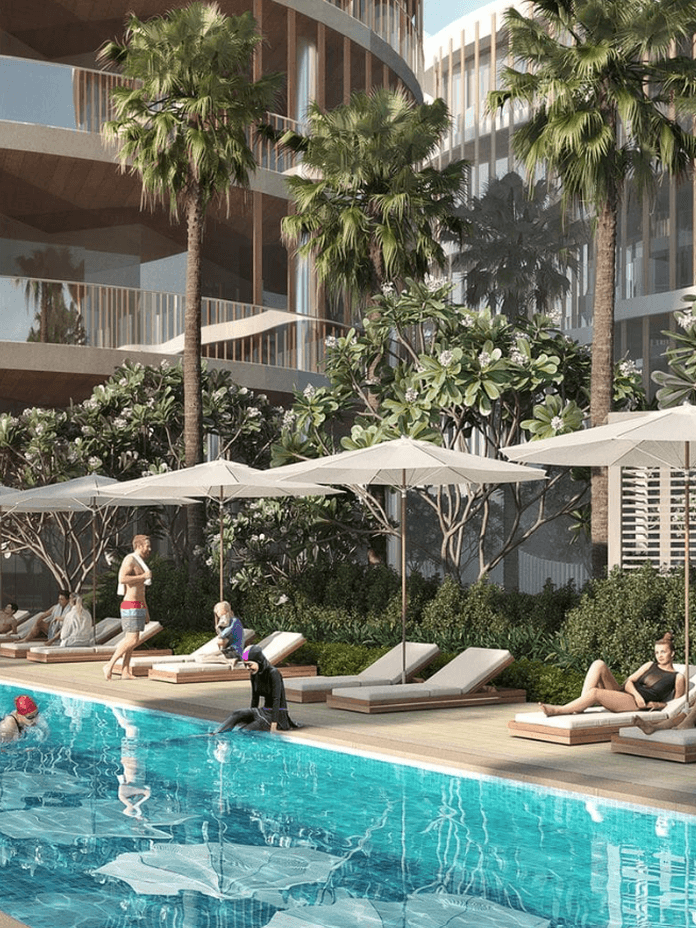 Modern apartment complex with a pool, surrounded by palm trees and lounge chairs. People relax under umbrellas on a sunny day.