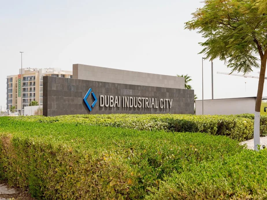 Sign reading "Dubai Industrial City" surrounded by green hedges, with buildings and a tree in the background.