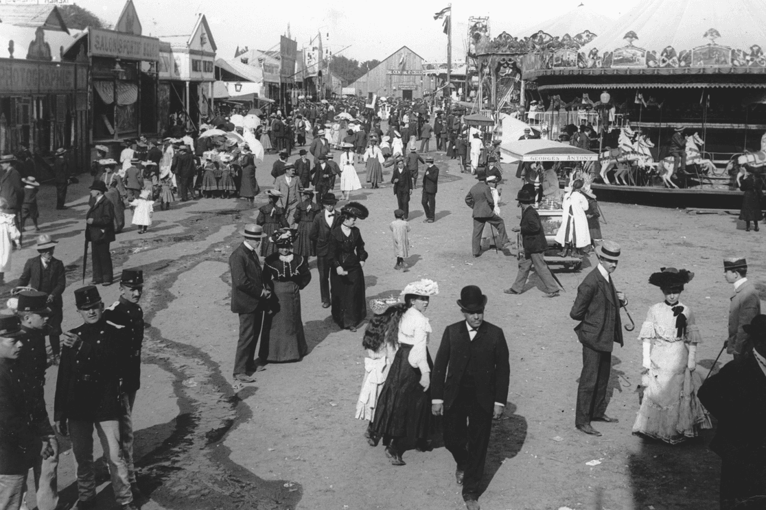 Vintage fair scene with people in early 20th-century attire walking along a dirt path, surrounded by shops and a carousel.