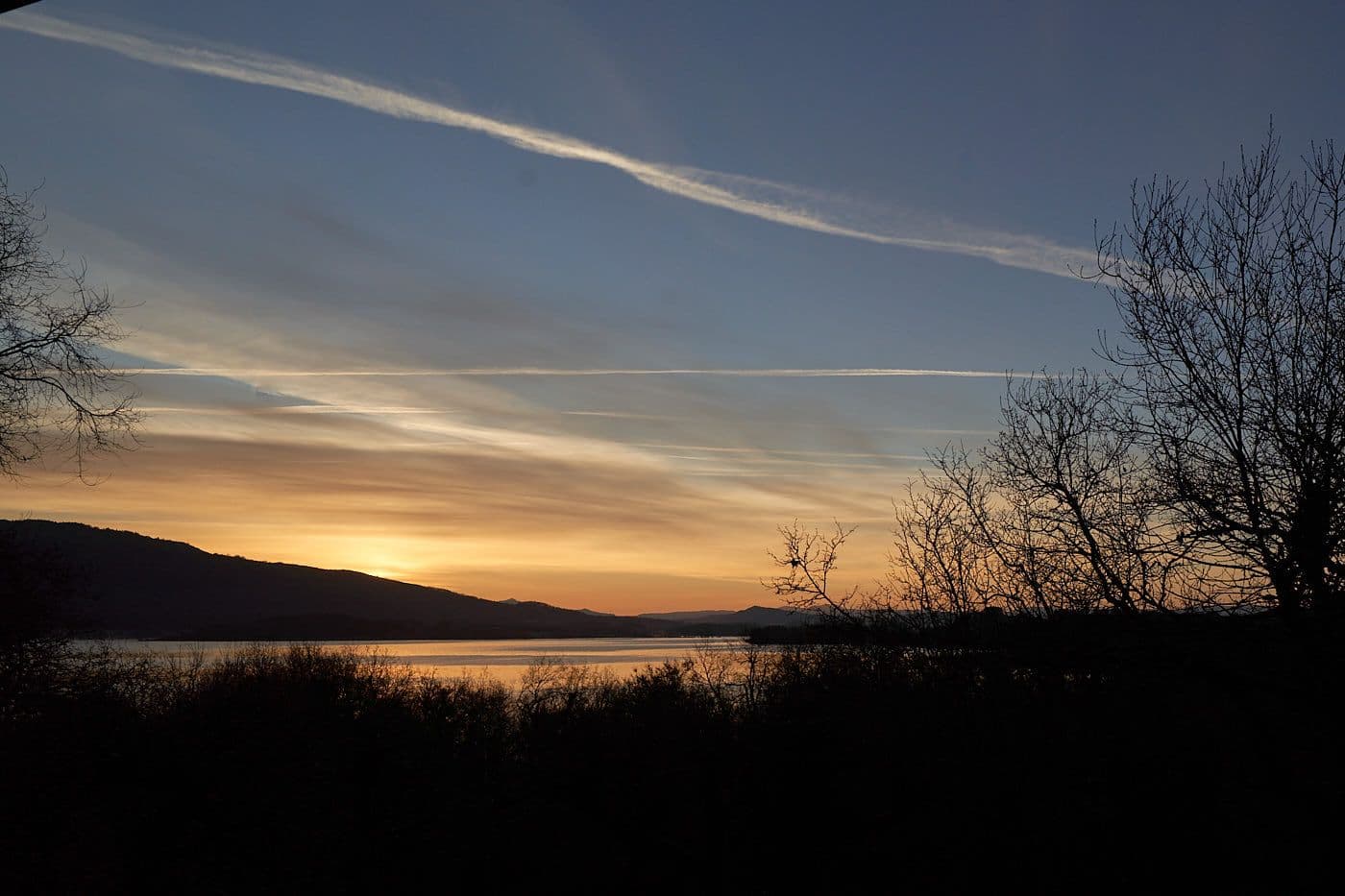Ein wunderschöner Sonnenuntergang über einem See, umgeben von silhouettierten Bäumen und Hügeln im Hintergrund. Der Himmel zeigt sanfte Farben von Orange bis Blau mit einigen Kondensstreifen